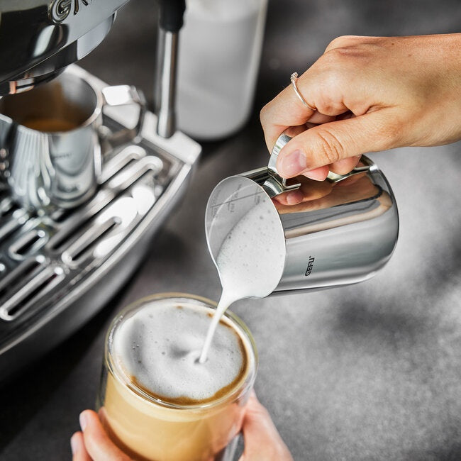 GEFU Melkkan Barista - 250 ml, hand die opgeschuimde melk vanuit de kan in een glas koffie schenkt bij het koffiezetapparaat