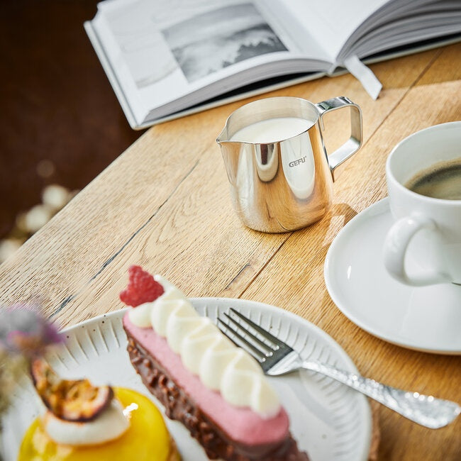 GEFU Melkkan Barista - 150 ml op een houten tafel naast een kop koffie en een bordje met gebak en fruit