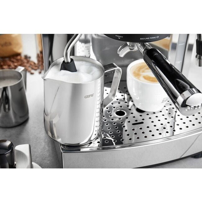 GEFU Melkkan Barista - 350 ml gevuld met melkschuim naast het espressoapparaat en een kop koffie met latte art