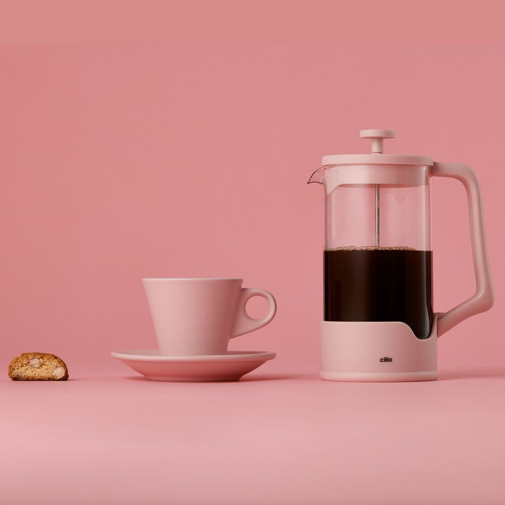 Roze cafetière met zwarte koffie naast een roze kop en schotel en een koekje met amandelen op roze achtergrond