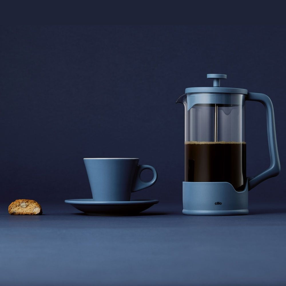 Blauwe cafetière gevuld met koffie naast een blauw kopje met schotel en een koekje voor een donkerblauwe achtergrond