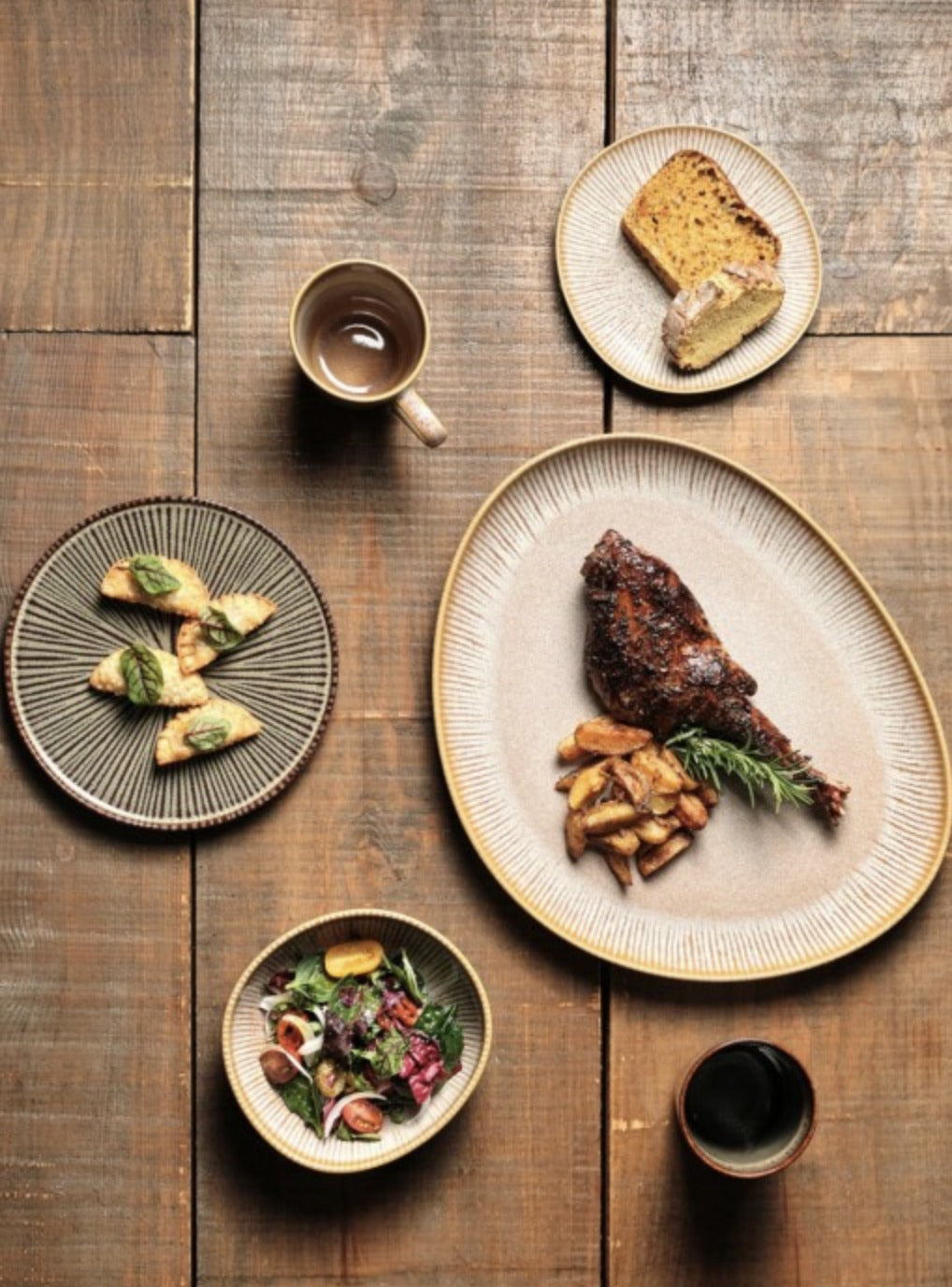 Gedekte houten tafel met een volledige Shangri-la tafelsetting: kopjes thee, een bord met cake, hapjes op een donkere ronde schaal, een ovale schaal met gebraden gerecht en een kom salade