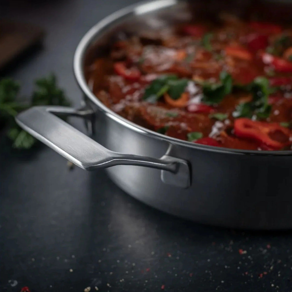 Close-up van de sauteerpan gevuld met een tomatenragout met paprika en verse kruiden, met het hulphandvat op de voorgrond.