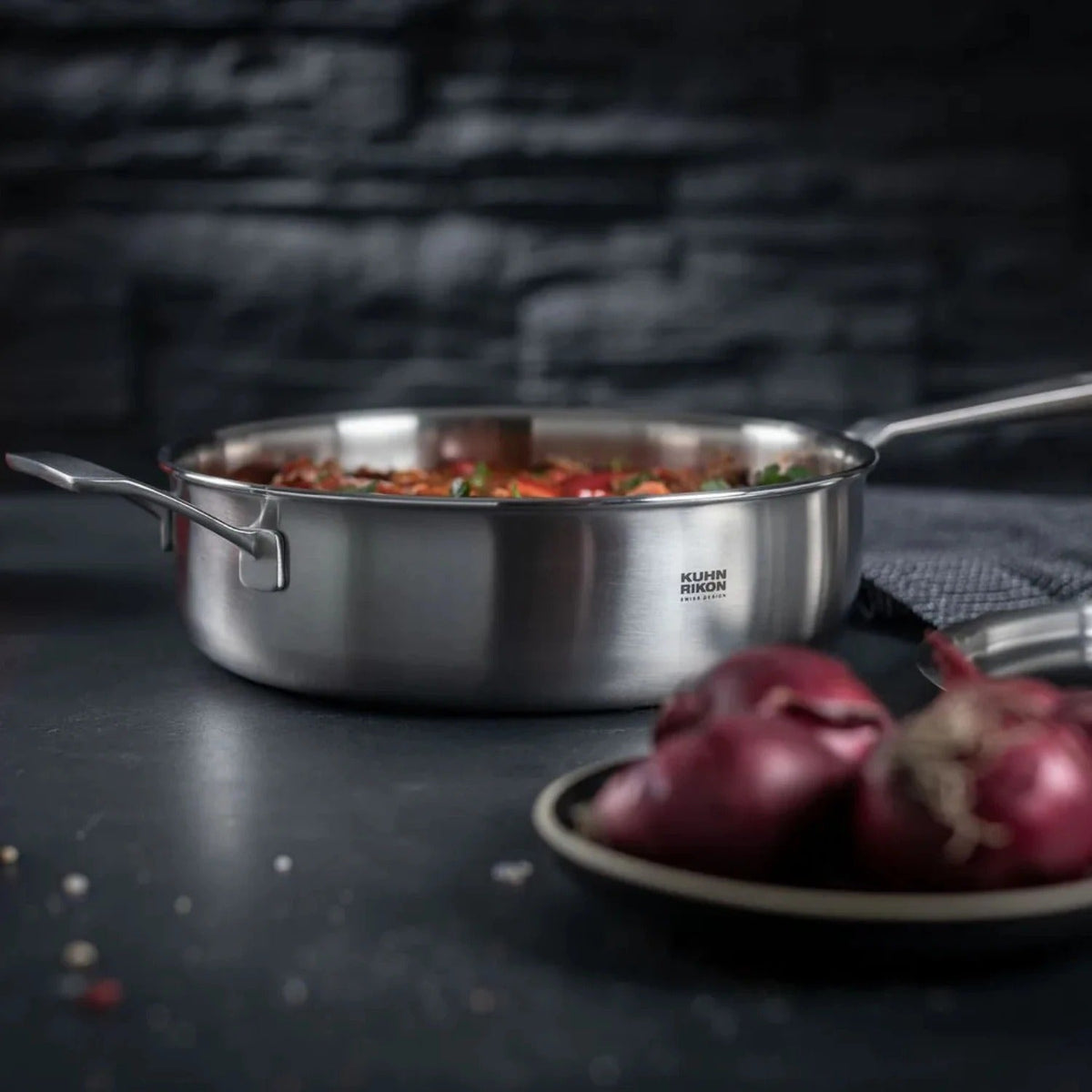 Kuhn Rikon Culinary 5-Ply sauteerpan 28 cm gevuld met een gebakken tomatenmengsel, op een donker aanrecht naast een bord rode uien.