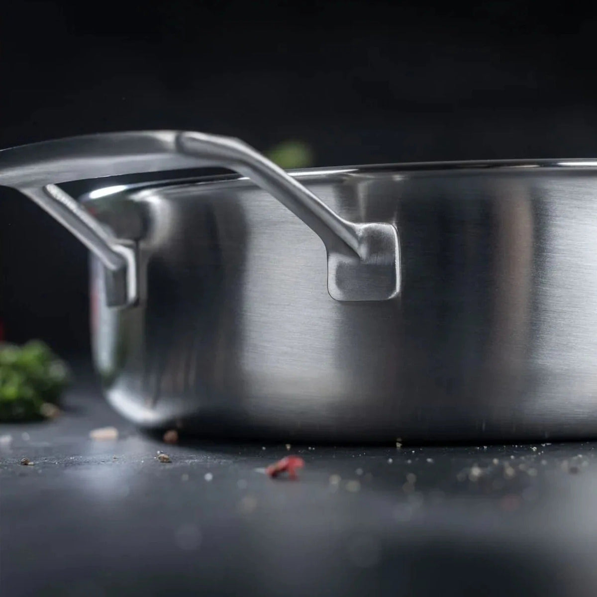 Close-up van het roestvrijstalen hulphandvat van de Kuhn Rikon sauteerpan 28 cm, met kruiden en een rode peper op de achtergrond.