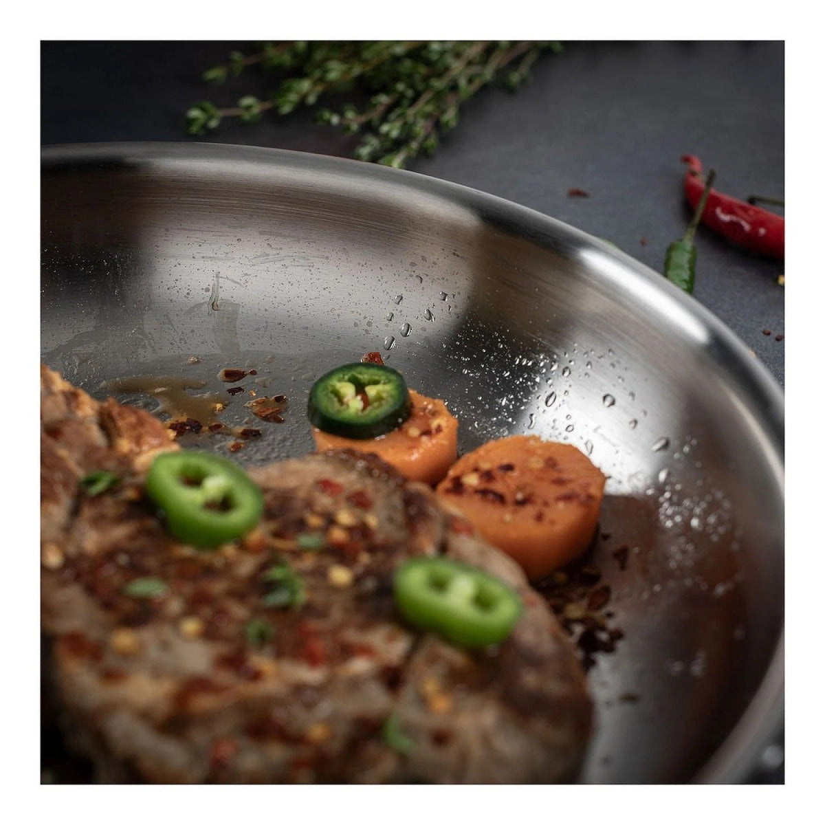 Close-up in de Culinary 5-Ply koekenpan 20 cm met gegrild vlees, plakjes jalapeño en stukjes wortel, met waterdruppels zichtbaar op het roestvrijstalen oppervlak.