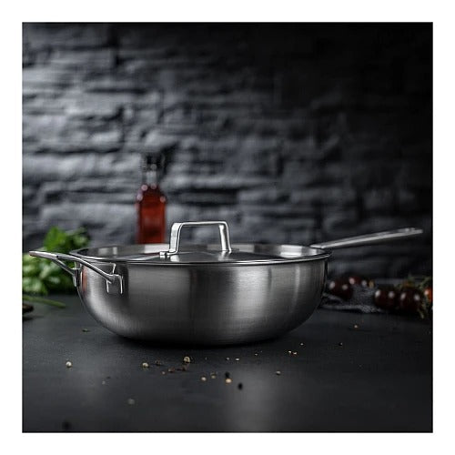 De Culinary 5-Ply wok met deksel 28 cm met de deksel erop op een donker aanrecht, met kruiden, cherrytomaten en een fles olie op de achtergrond.