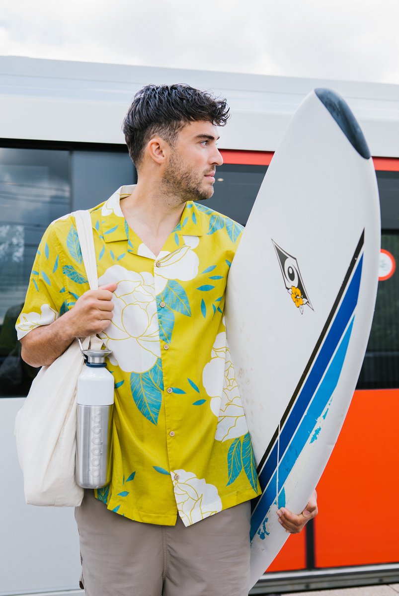 Man met een surfboard onder de arm heeft de Dopper Steel - 1,1 liter aan een tas bevestigd, met een tram op de achtergrond