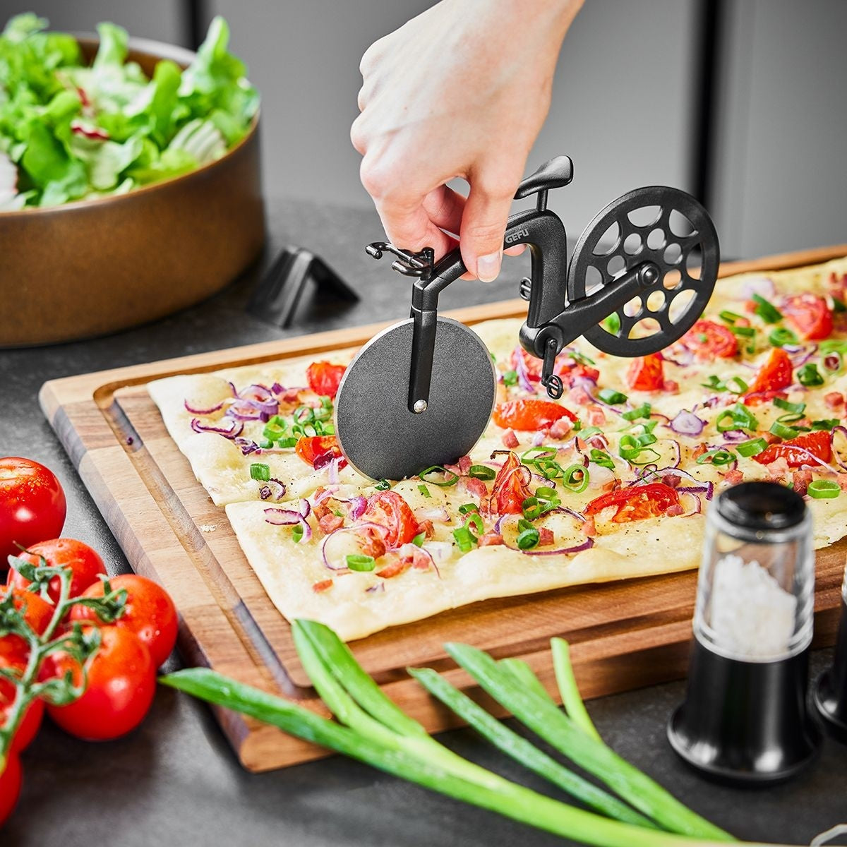 GEFU Pizzasnijder Farado, een hand snijdt met de fietsvormige pizzasnijder door een pizza met tomaat en rode ui op een houten plank, met een kom sla en verse tomaten ernaast