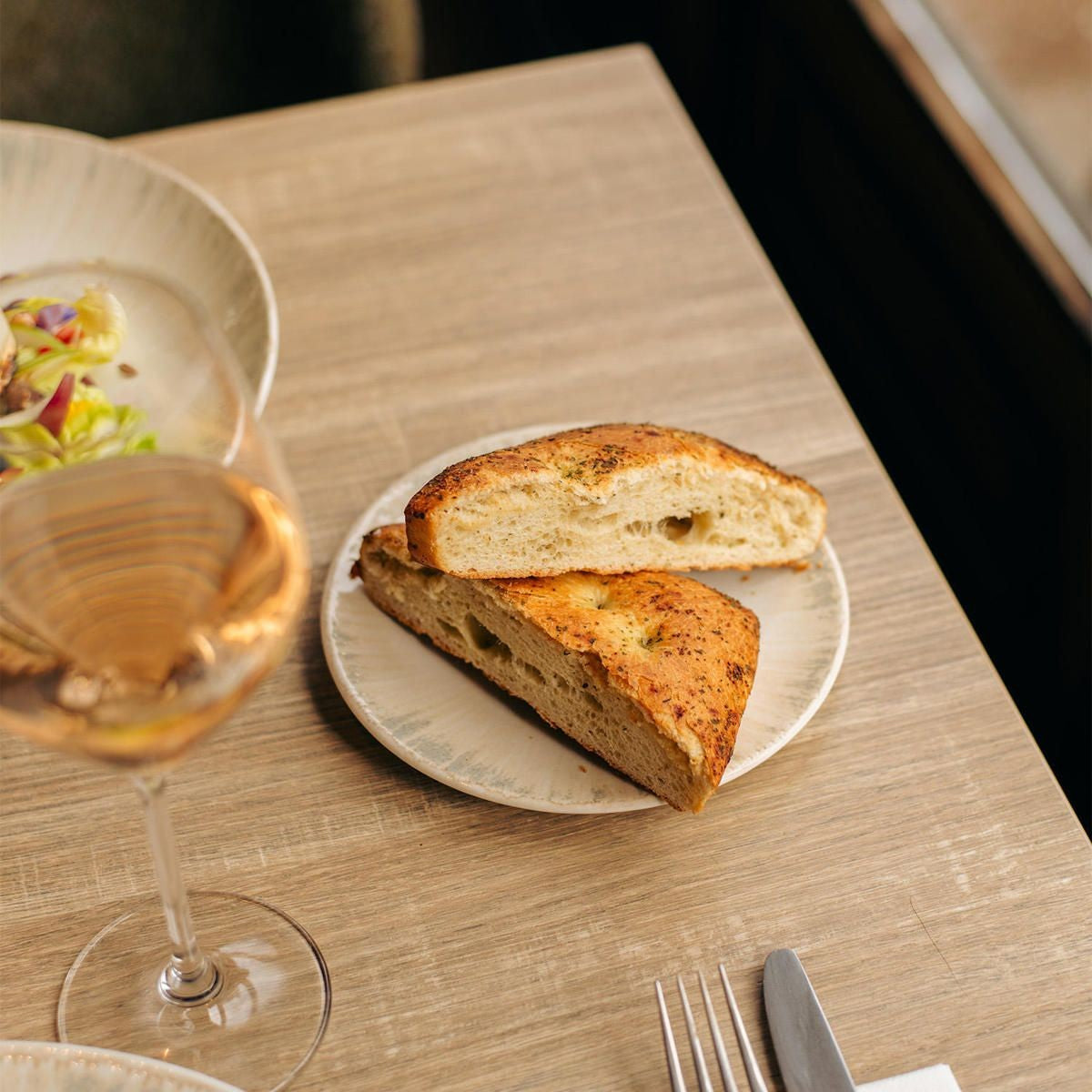 Twee stukken focaccia op het ivoorkleurige bord op een houten tafel, naast een wijnglas, bestek en een saladekom