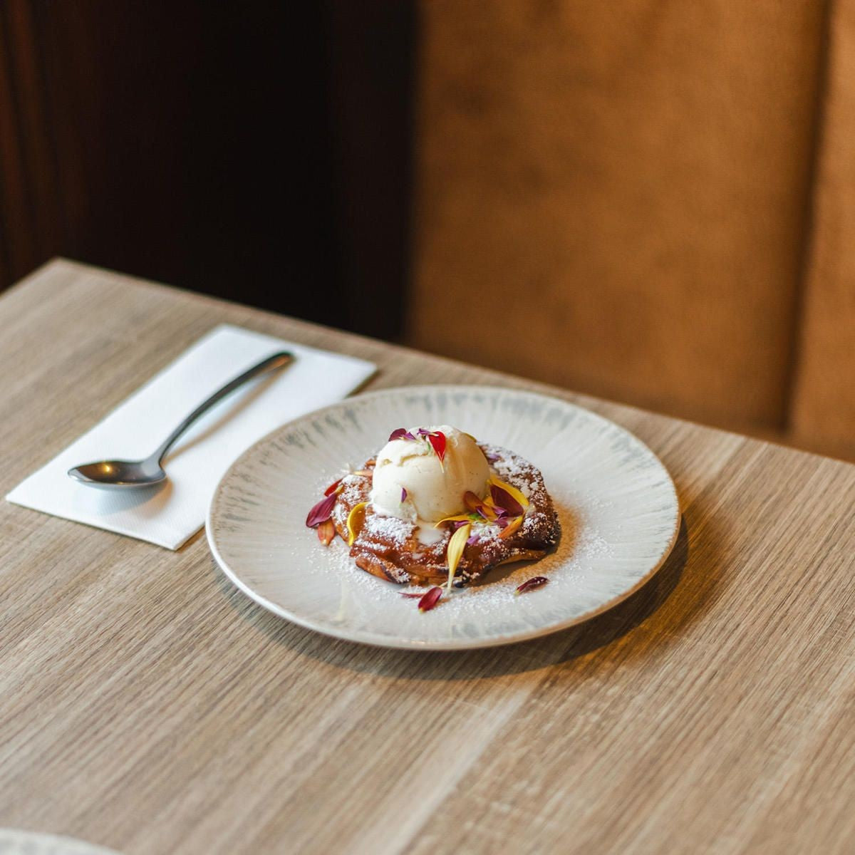 Bord met een bolletje vanille-ijs op een gebakken dessert met bloemblaadjes, naast een lepel op een servet op houten tafel
