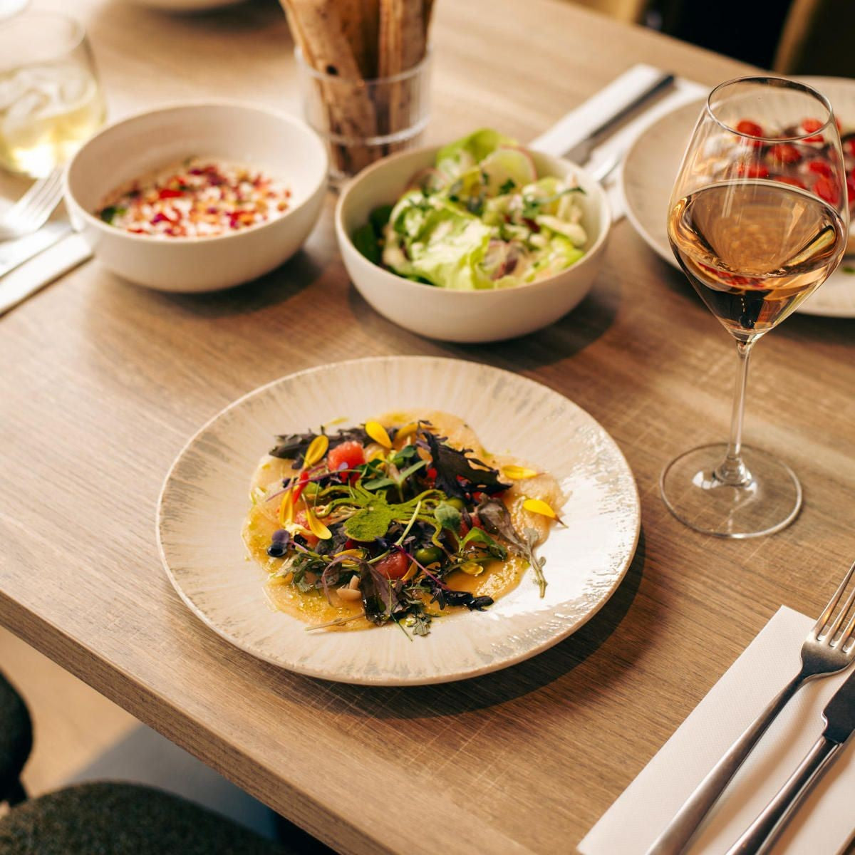 Bord met carpaccio, kruidenmengsel en eetbare bloemen op een houten tafel, naast salade en wijnglas