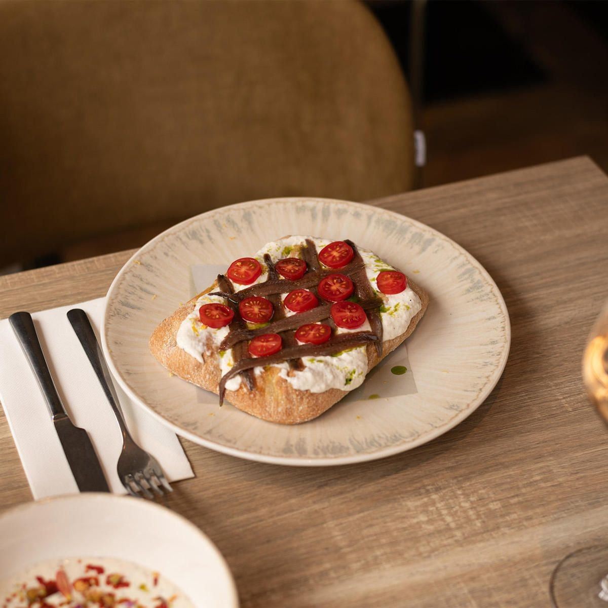 Schuin geplaatst bord met een stuk boerenbrood met ricotta, ansjovis en tomaatjes, naast mes en vork op een servet
