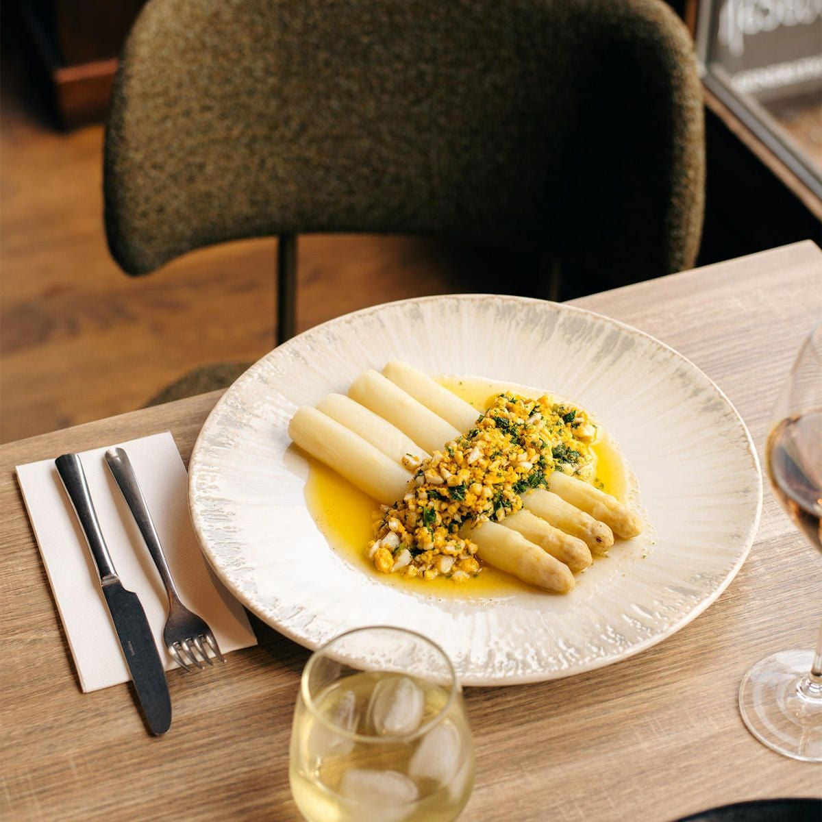 Bord met witte asperges, botersaus en gehakt ei en peterselie, naast bestek op servet en twee glazen op tafel