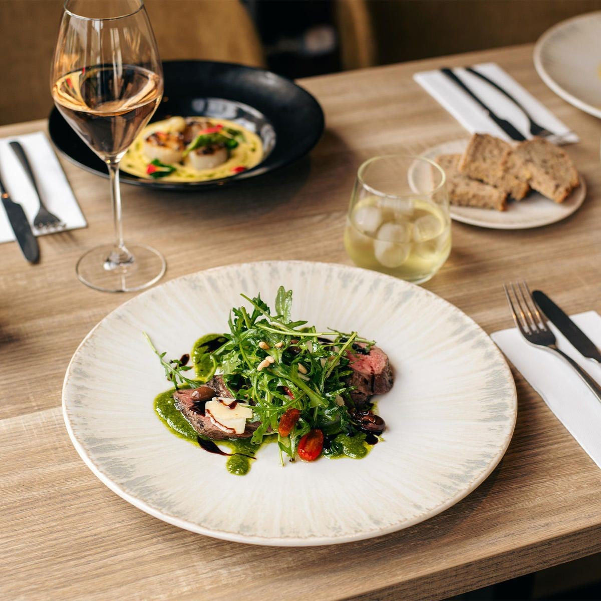 Groot bord met plakjes gebakken vlees, rucola, pijnboompitten, pesto en balsamico, naast een wijnglas en een zwart bord