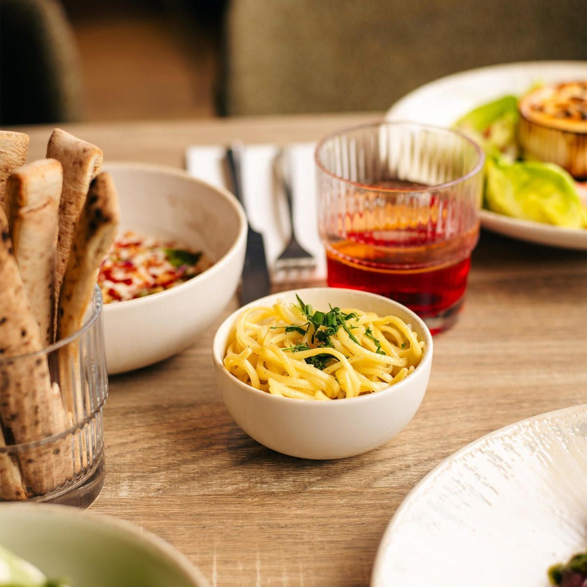 Kleine ivoorwitte kom gevuld met spaghetti en kruiden op houten tafel naast brood in een gestreept glas