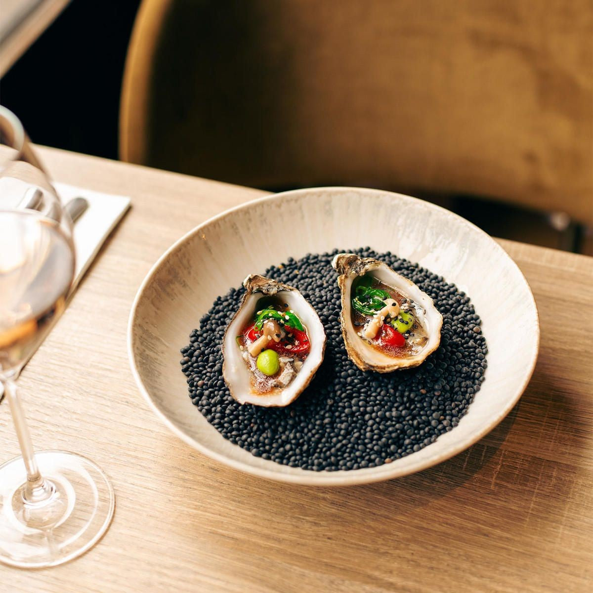 Twee oesters met garnering op zwarte linzen in de brede kom, op houten tafel naast een wijnglas