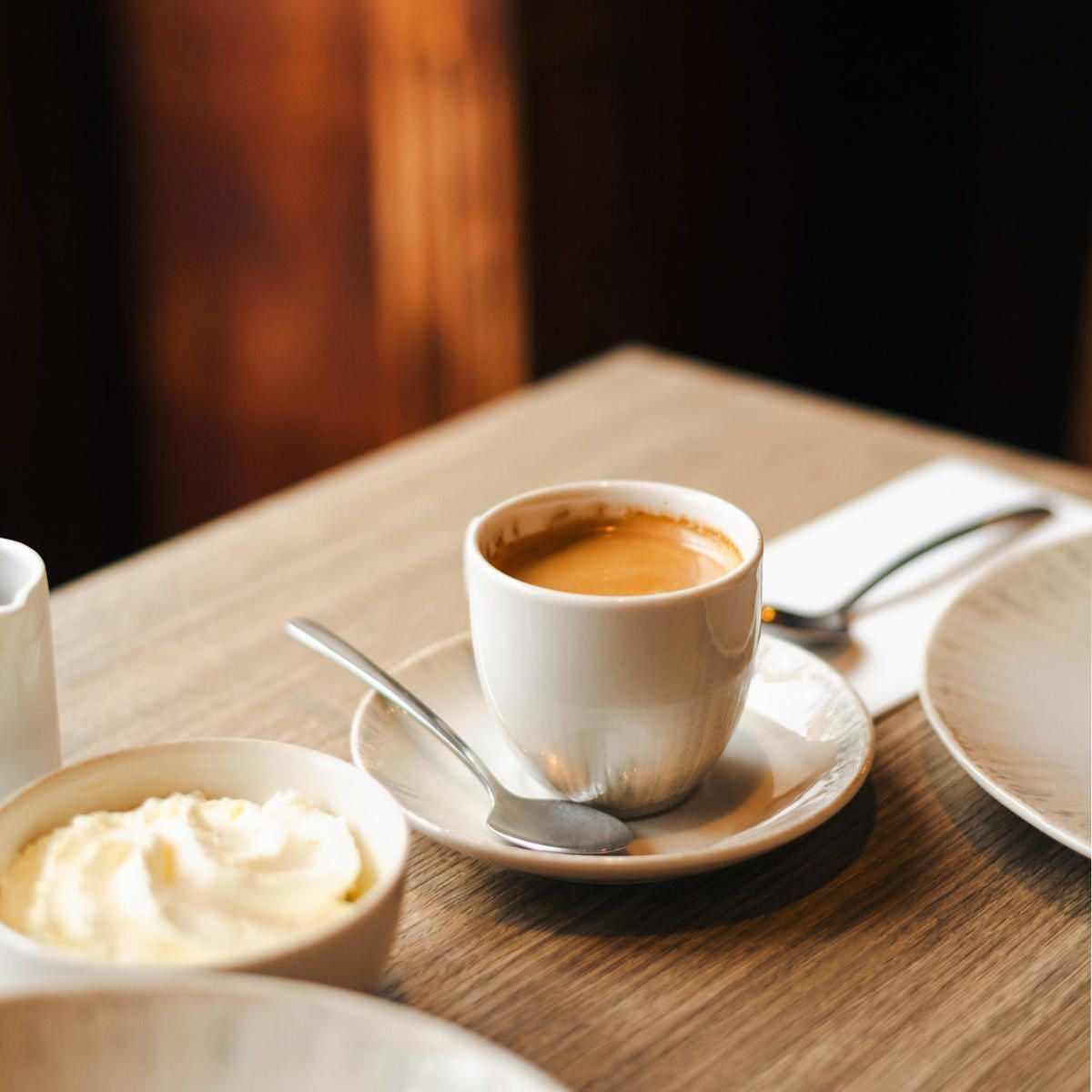 Espresso met crema in een ivoorwitte kop op een schotel met lepel, naast een schaaltje met slagroom