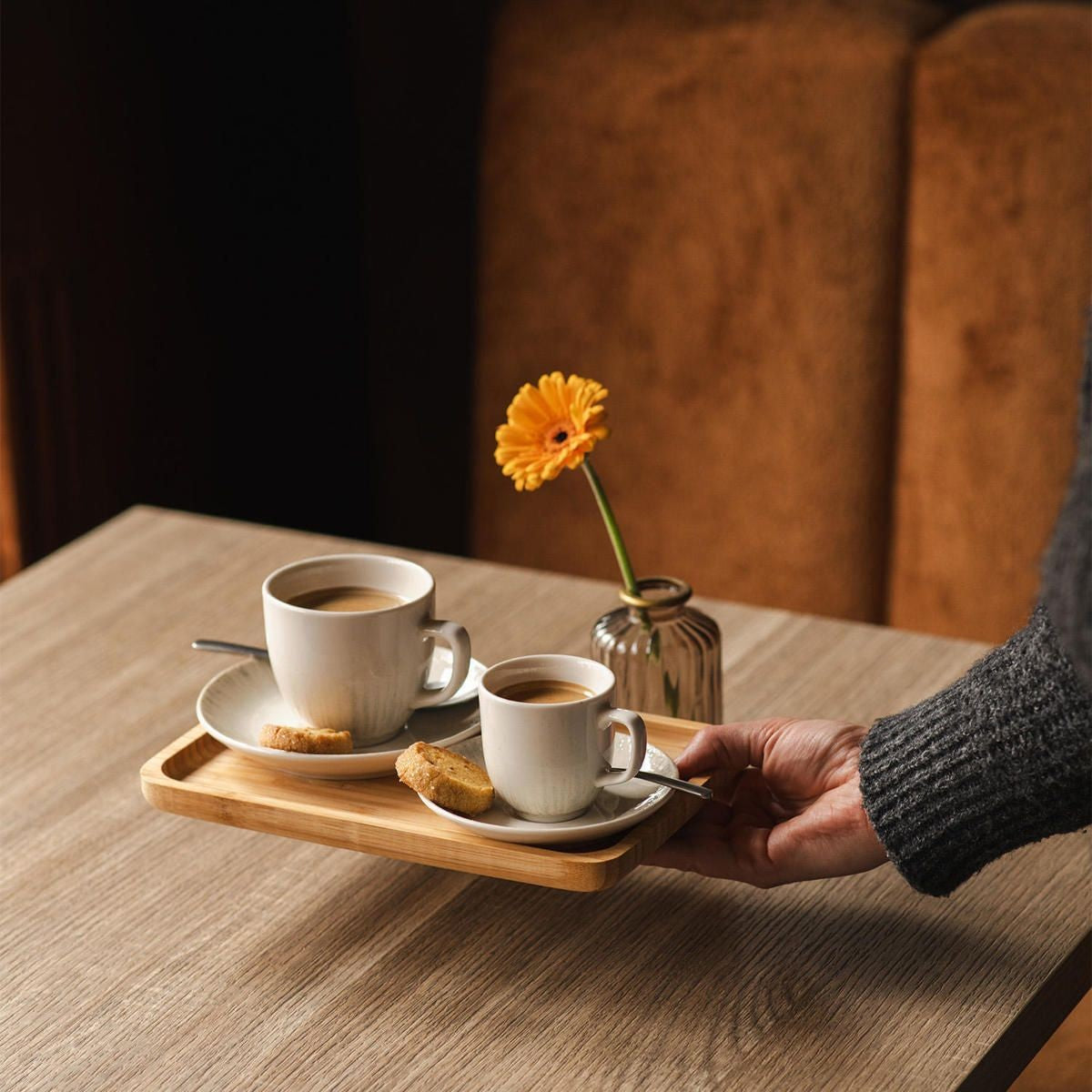 Hand draagt een houten dienblad met een grote en een kleine kop op schotels, koekje en een gele gerbera