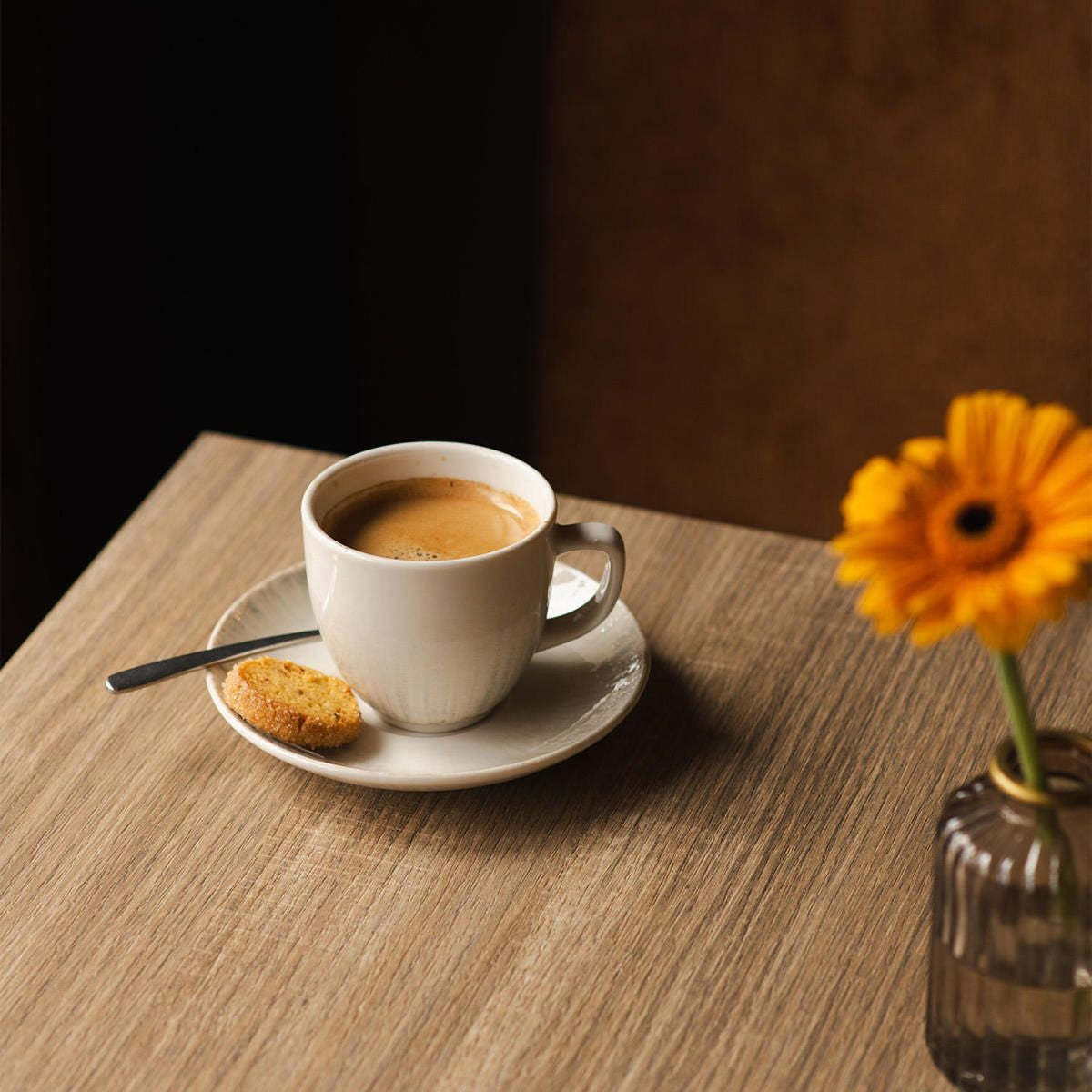 Kop met koffie op een schotel met lepel en een koekje, naast een oranje gerbera op een houten tafel