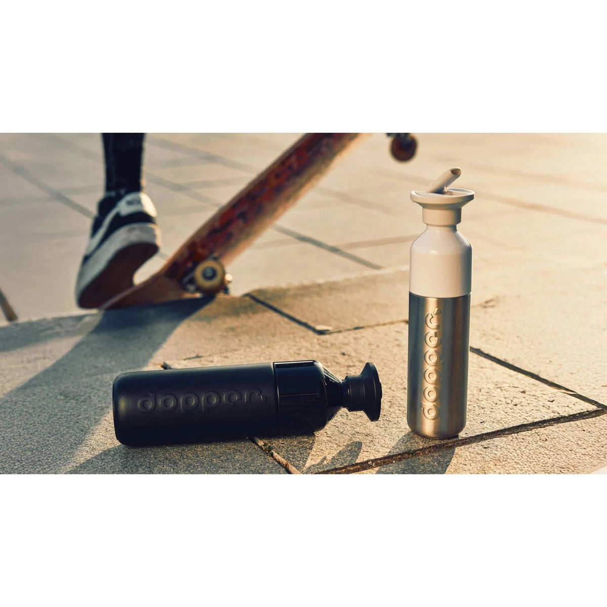 Een zilverkleurige en een zwarte Dopper Steel x Flip Straw op straat bij een skatepark, met een skateboarder op de achtergrond