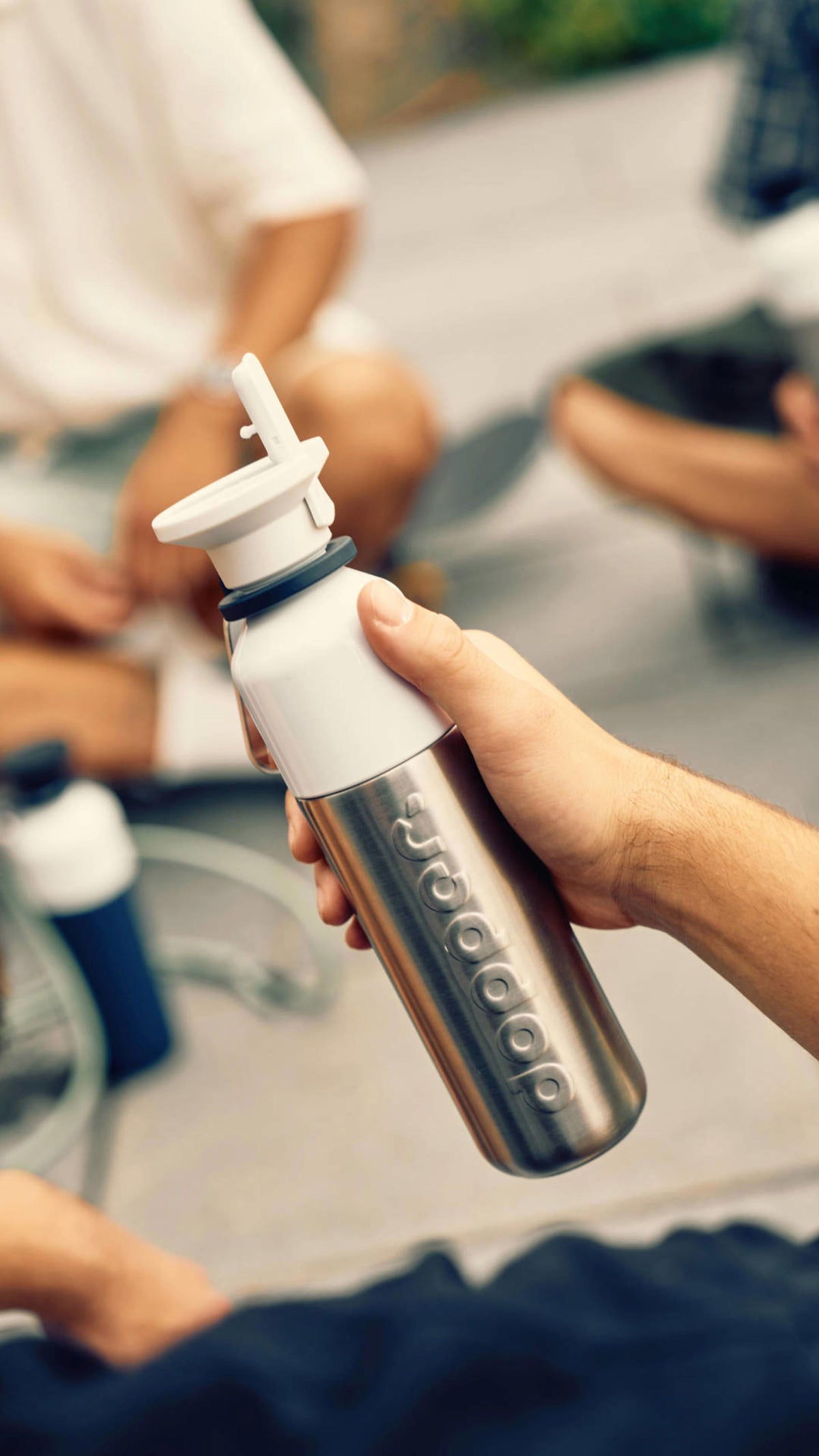 Hand houdt de Dopper Steel x Flip Straw vast met de dop opengeklapt, temidden van een groep vrienden buiten