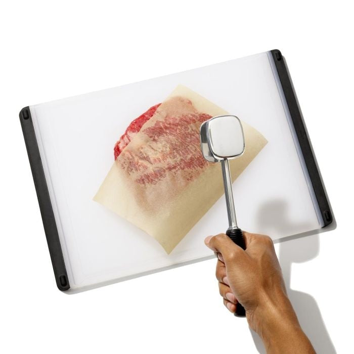 OXO Good Grips Vleeshamer Aluminium, in de hand vastgehouden boven stuk vlees tussen bakpapier op snijplank