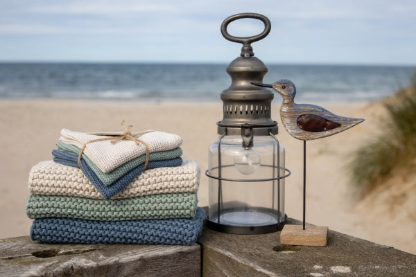 Solwang Design Vaatdoekjes set van 3 - Nordic Breeze lifestylefoto op een houten balk aan het strand: de doekjes in crème, groen en blauw liggen gestapeld en samengebonden met touw naast een metalen lantaarn met glazen kap en een houten vogelfiguurtje, met duinen en zee op de achtergrond.