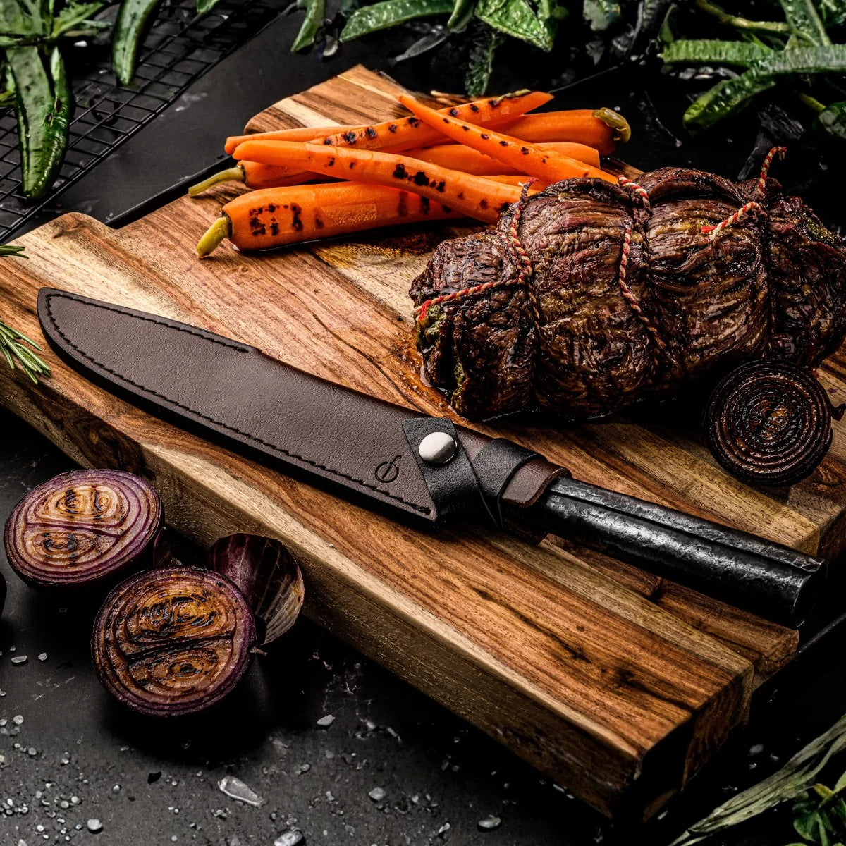 Vleesmes met leren hoes op een houten plank naast gegrilde rollade en groenten