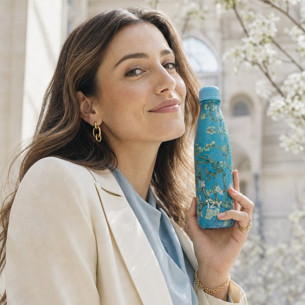 Vrouw houdt de turquoise IZY-thermosfles met Amandelbloesem van Van Gogh vast bij een bloesemboom
