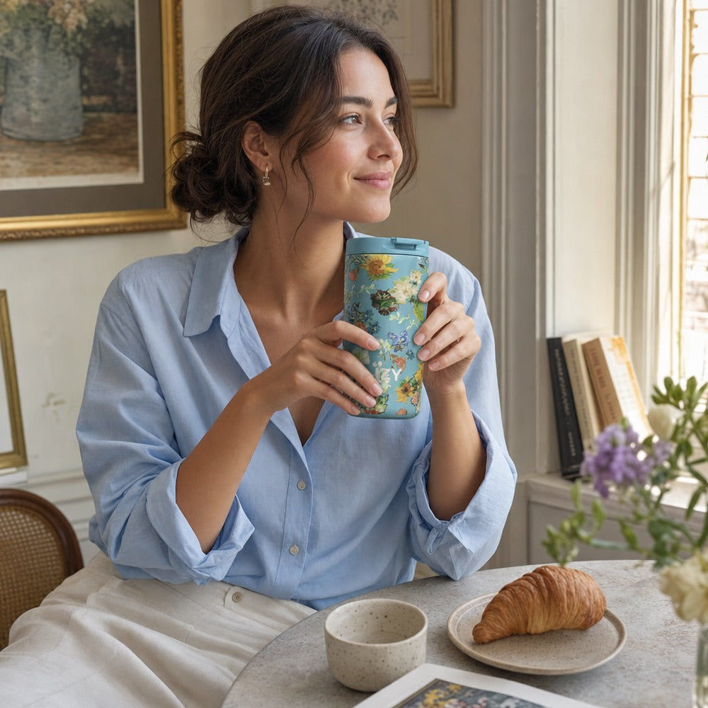 Vrouw houdt de IZY Thermosbeker Vincent's Flowers vast aan tafel met een croissant
