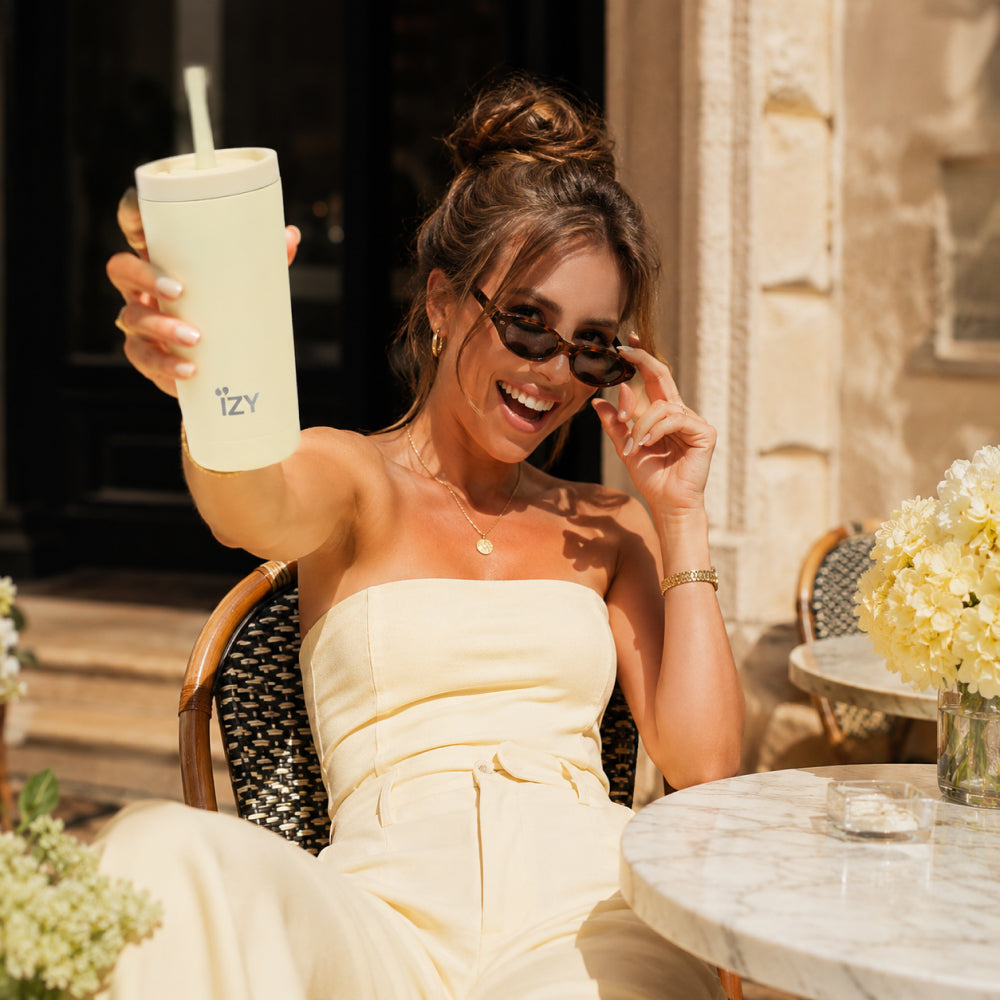 Vrouw met zonnebril houdt de lichtgele IZY Tumbler Butter Yellow vast aan een terrastafel