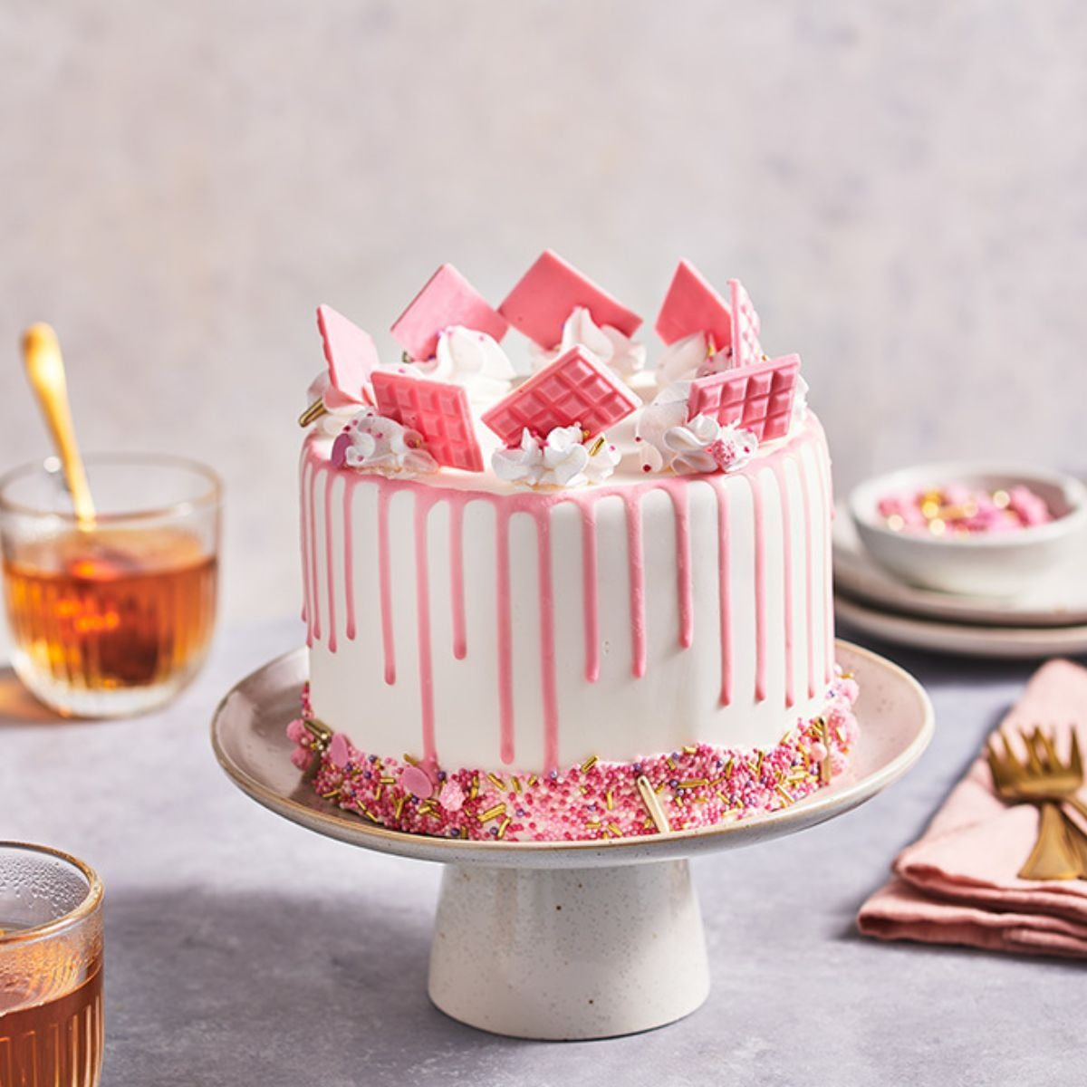 Drip cake met roze ganache en roze chocoladereepjes op een taartplateau, met een glas thee en bestek op tafel