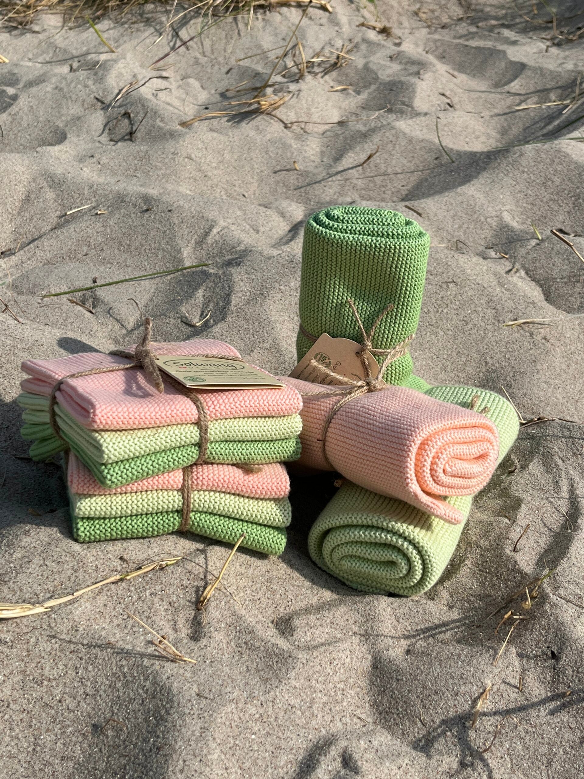 Solwang Design Vaatdoekjes set van 3 - Papaya Combi lifestylefoto op zand: meerdere doekjes in zalmroze en groen liggen gestapeld en samengebonden met touw, met daarnaast een los opgerold groen en een los opgerold zalmroze doekje, voorzien van het kraftpapieren Solwang-label.