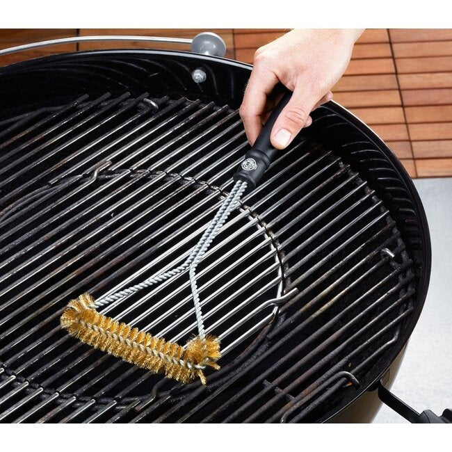 GEFU Driehoekige Barbecueborstel, een hand die de borstel met de messing haren gebruikt om het rooster van een kogelbarbecue schoon te maken