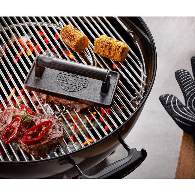 GEFU Vleesgewicht op de grill gebruikt om vlees plat te drukken naast maiskolven, met een ovenwant op de achtergrond