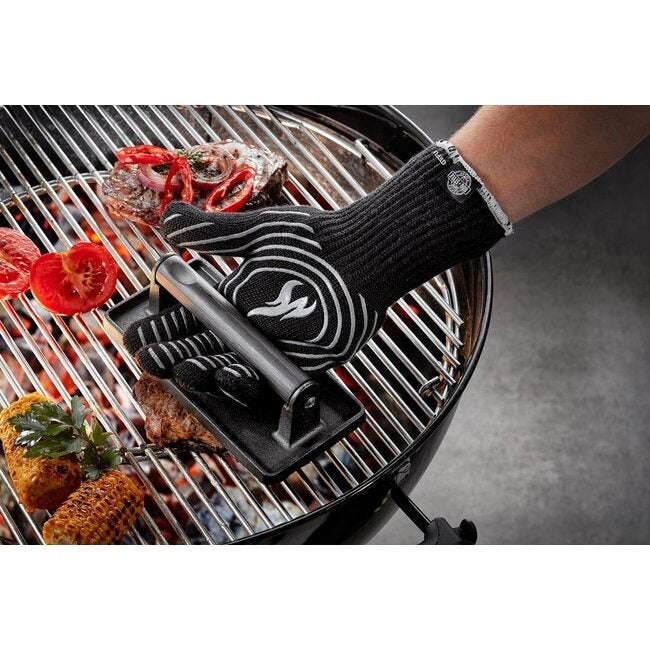 GEFU Barbecuehandschoen aan een hand die met een tang over een brandende barbecue reikt met vlees, maiskolven, tomaten en citroen op het rooster