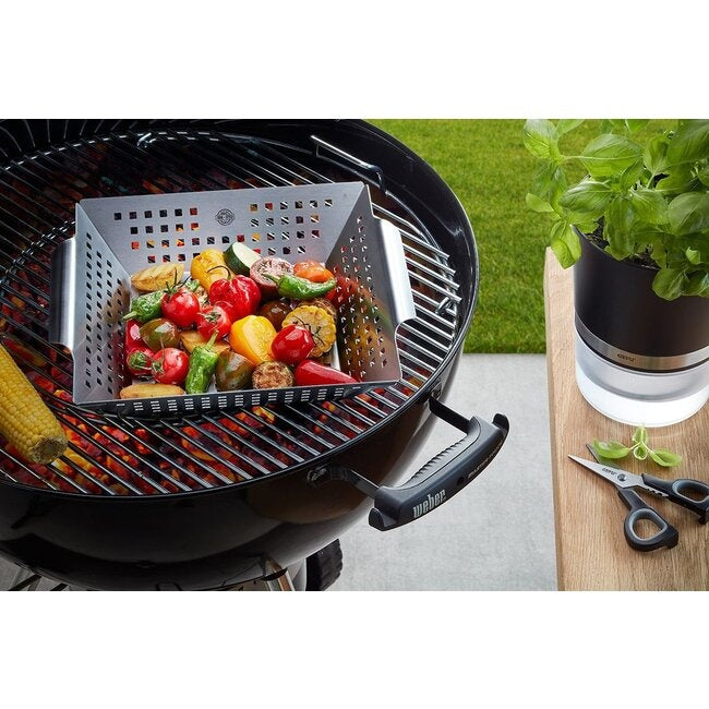 GEFU Grillplaat - Groot, gevuld met gegrilde paprika en maiskolven op het rooster van een kogelbarbecue met gloeiende kolen, met een basilicumplant en schaar op een houten tafel ernaast