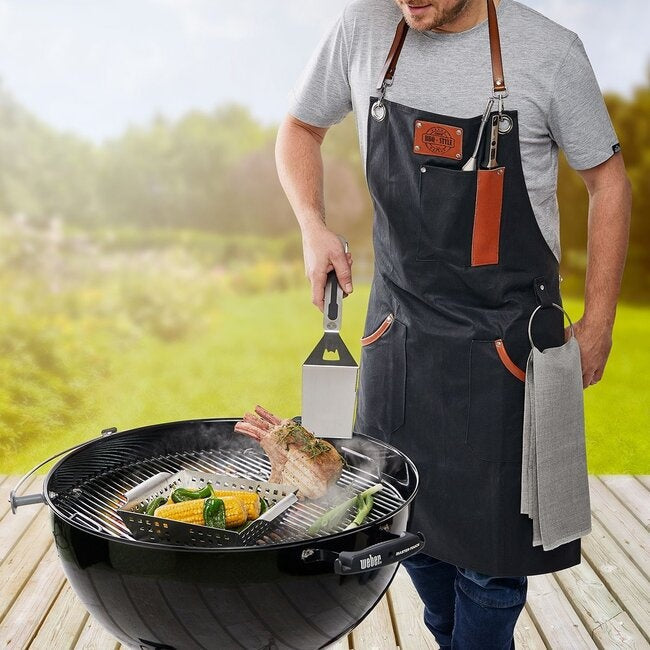 GEFU Barbecueschort gedragen door een man die bij een ronde barbecue staat met vlees en mais op het rooster, buiten