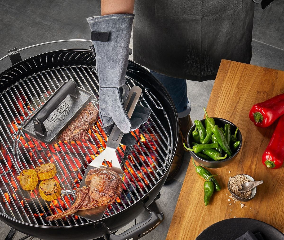 GEFU Barbecuehandschoenen - Suède Leer aan een hand die met een tang een groot stuk vlees keert boven gloeiende kolen, met maiskolven, pepers en een houten plank ernaast