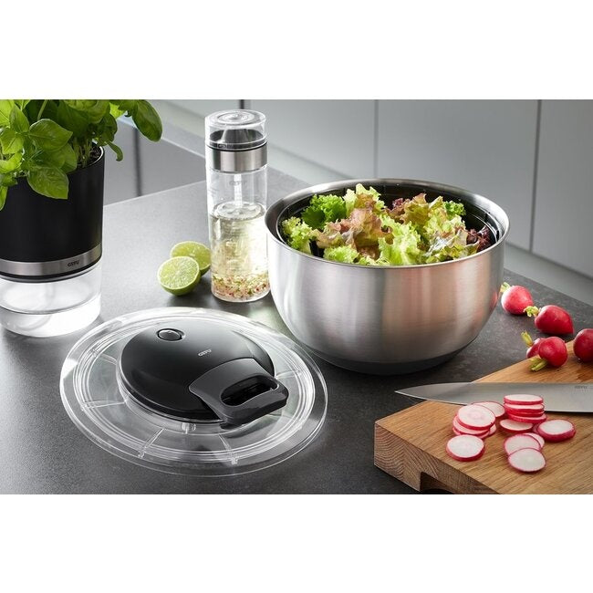 GEFU Saladespinner Pullit RVS met de losse deksel naast de RVS kom gevuld met slablaadjes, kruiden, limoen en radijs, met een mes en gesneden radijsschijfjes op een houten plank