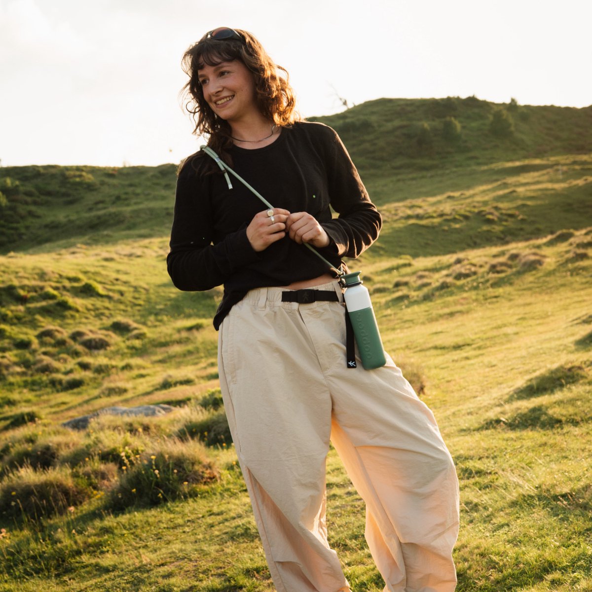 Vrouw wandelt in de heuvels bij zonsondergang met de Dopper Insulated Pine Green aan een koord om haar nek