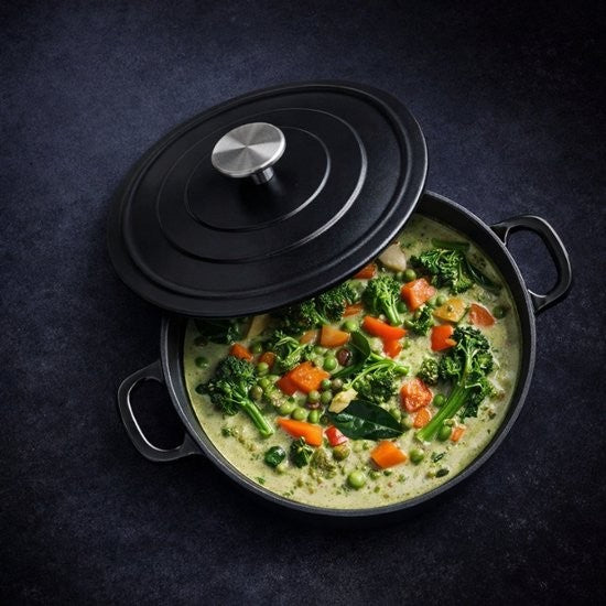 Royal Van Kempen braadpan gevuld met een groene curry met broccoli, doperwten en wortel, het deksel leunt schuin tegen de pan op een donkere ondergrond.