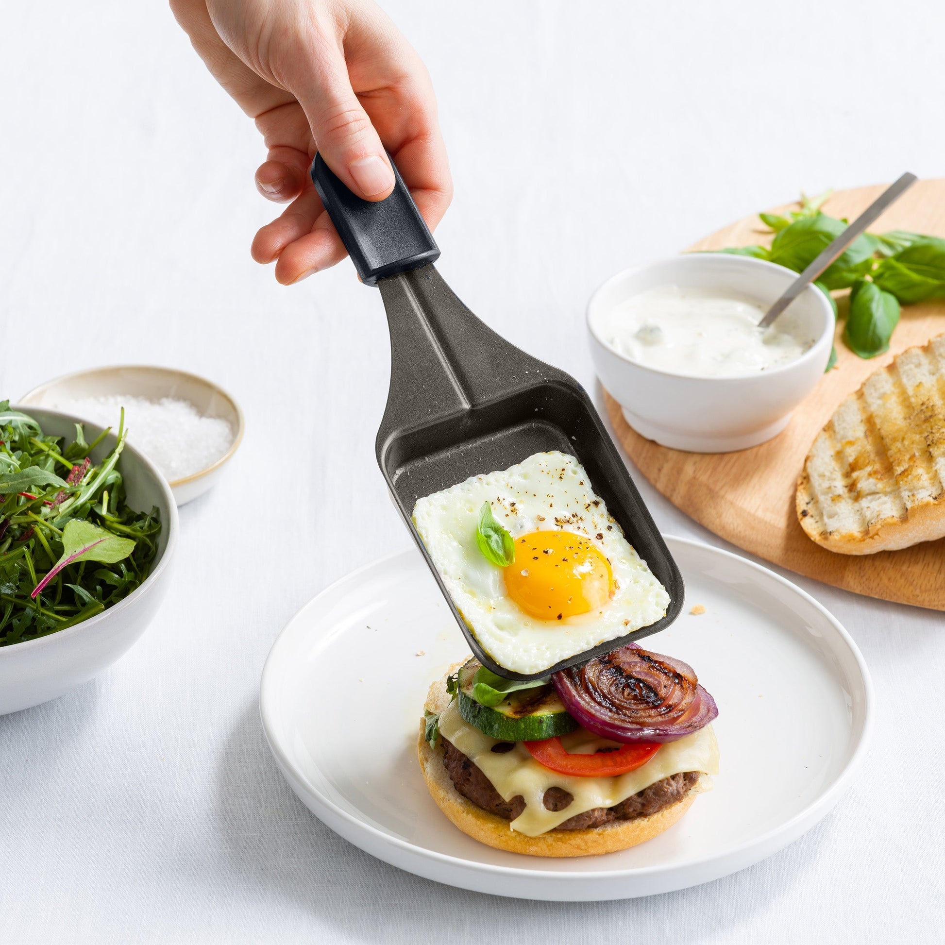Hand houdt een raclettepannetje met spiegelei boven een burger, van de BK Connect raclette.