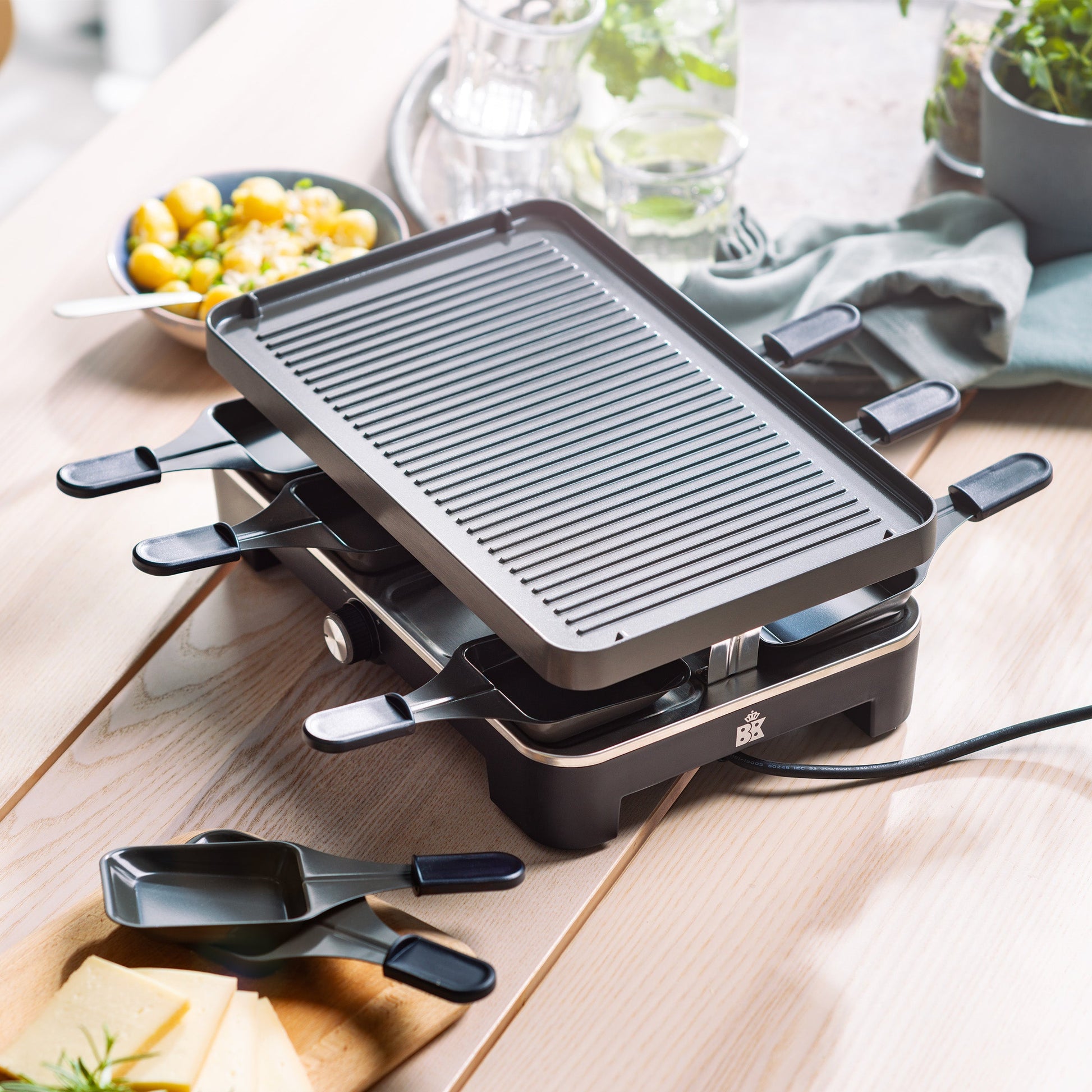 Zwarte BK Connect raclette met grillplaat en pannetjes op tafel, kaas ligt ernaast.