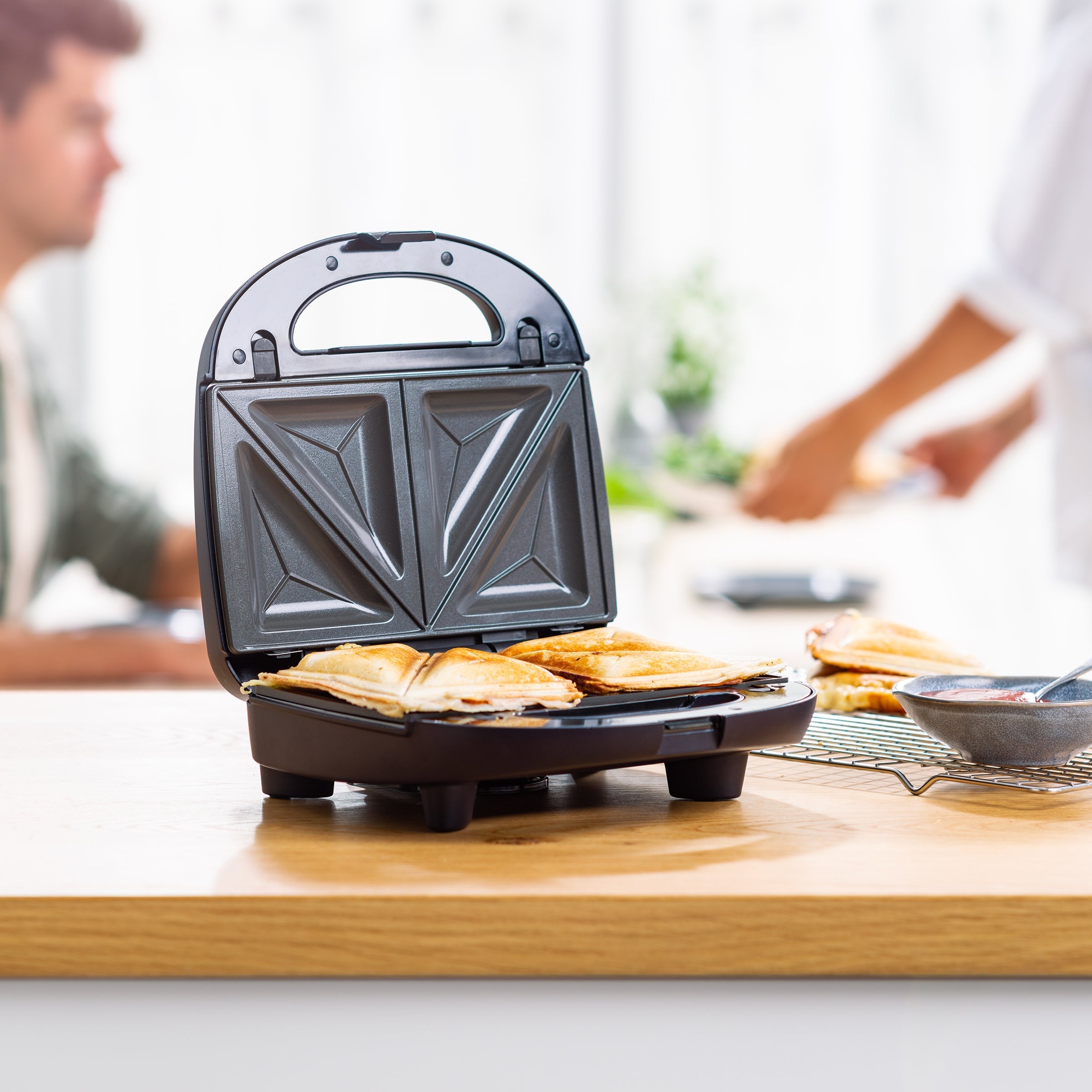 Zwarte BK Connect grill geopend met tosti's erin, twee personen op de achtergrond.