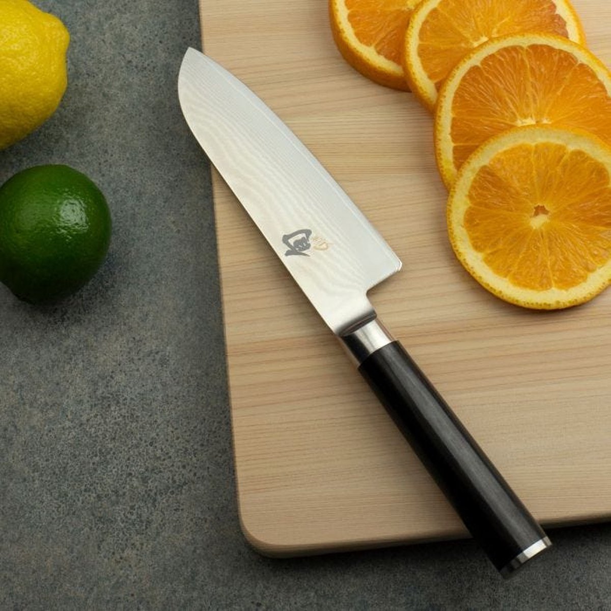 Kai Shun Classic Santoku First Touch - 14 cm, op een houten snijplank naast uitgesneden sinaasappel-, limoen- en citroenschijfjes