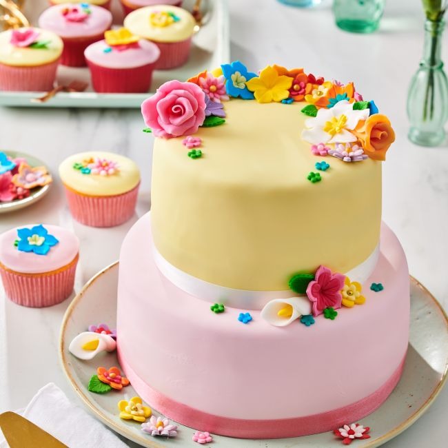 Taart met twee lagen in geel en roze fondant en suikerbloemen naast cupcakes met bloemdecoratie