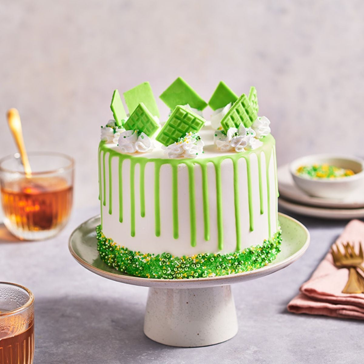 Drip cake met groene ganache en groene chocoladereepjes op een taartplateau, met een glas thee en bestek op tafel