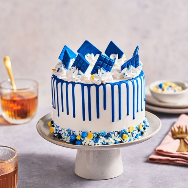 Taart met blauwe drip, blauwe chocoladeplakken en sprinkles onderaan op een taartplateau